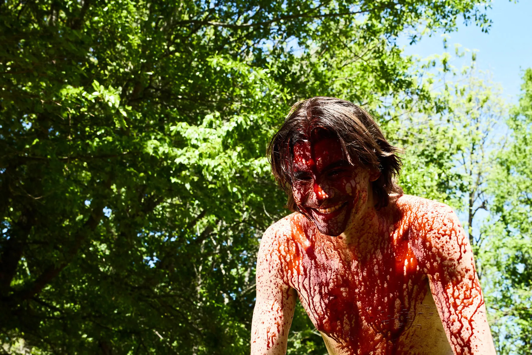 Andrew on the set of No Love Lost, shirtless and covered in stage blood, grinning, sunlit trees behind him